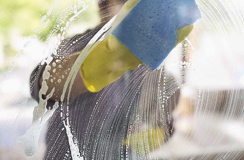 Een persoon wast een glazen venster met zeepachtig water en een blauwe spons, waarbij gele rubberen handschoenen worden gedragen.