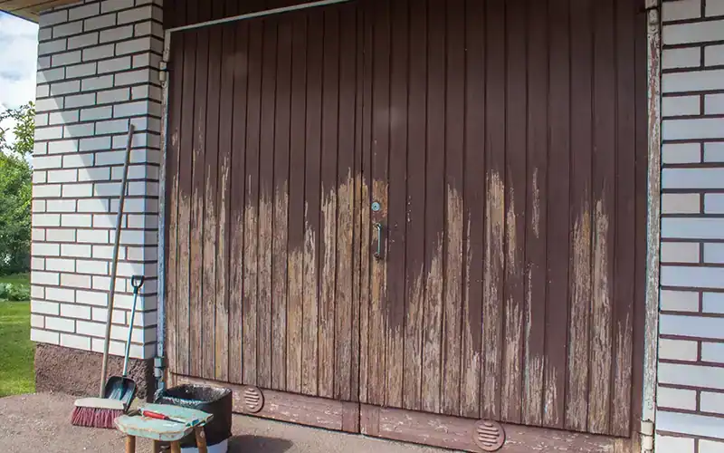 Garagedeuren van hout met gedeeltelijk afgebladderde verf en voorbereide schoonmaakbenodigdheden ernaast.
